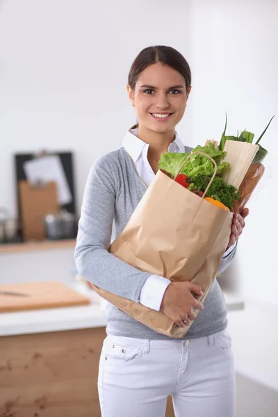 Wanita muda memegang tas belanja dengan sayuran berdiri di dapur. — Stok Foto