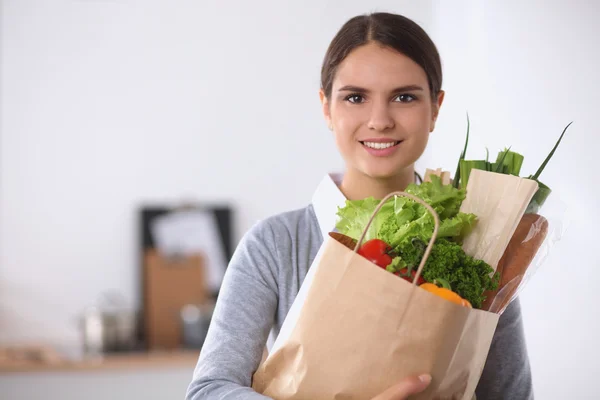 Mujer joven sosteniendo bolsa de la compra de comestibles con verduras de pie en la cocina. — Foto de Stock