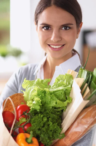 Mujer joven sosteniendo bolsa de la compra de comestibles con verduras de pie en la cocina. — Foto de Stock