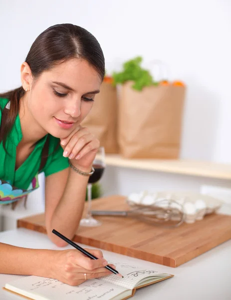 Happy beautiful woman standing in her kitchen writing on a notebook at home — Stock Photo, Image