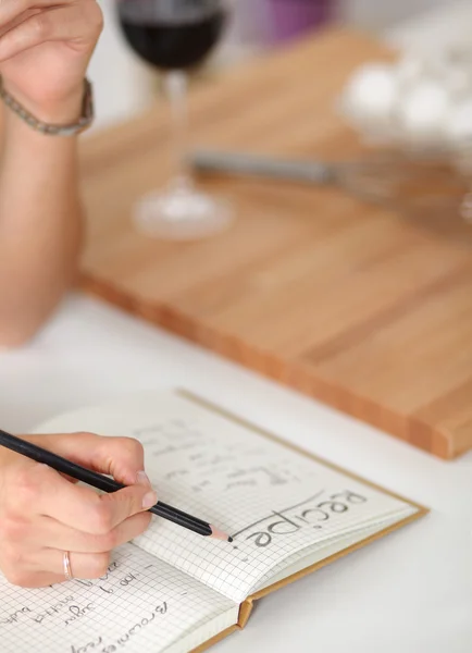 Happy beautiful woman standing in her kitchen writing on a notebook at home — Stock Photo, Image