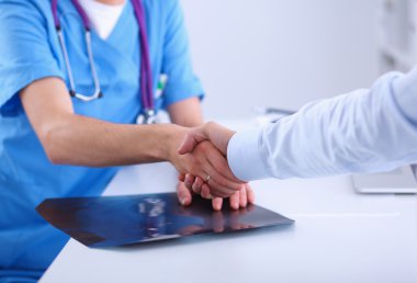 Doktor bir hastayla el sıkışıyor, ofiste masanın üzerinde oturuyor.