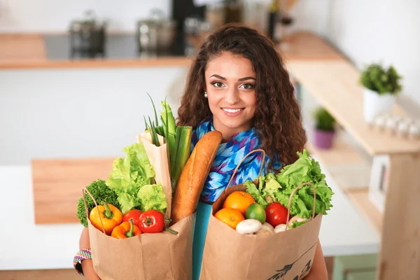 Mujer joven sosteniendo bolsa de la compra de comestibles con verduras de pie en la cocina. — Foto de Stock