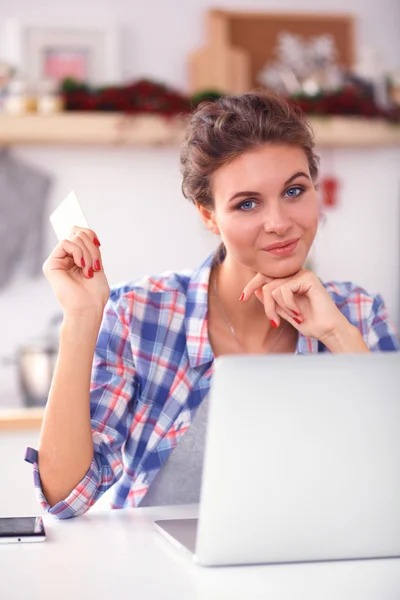 Mujer sonriente compras en línea utilizando la computadora y la tarjeta de crédito en la cocina —  Fotos de Stock