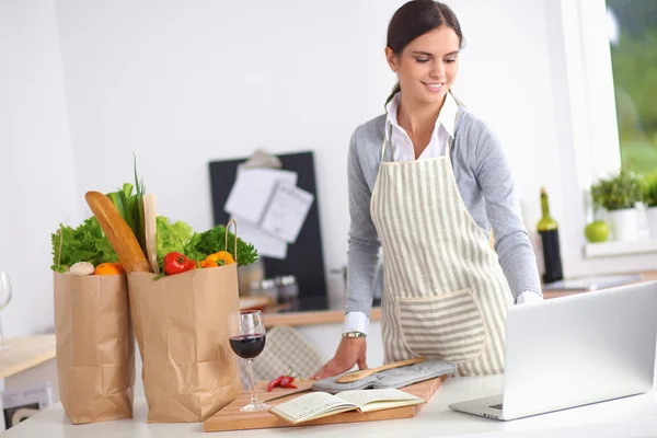 Beautiful young woman cooking looking at laptop screen with receipt in the kitchen — Stock Photo, Image