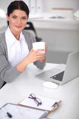 Genç bir iş kadını ofisinde fincanla masada oturuyor.