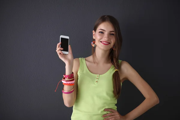 Hermosa mujer joven sosteniendo el teléfono celular, de pie cerca de la pared oscura — Foto de Stock