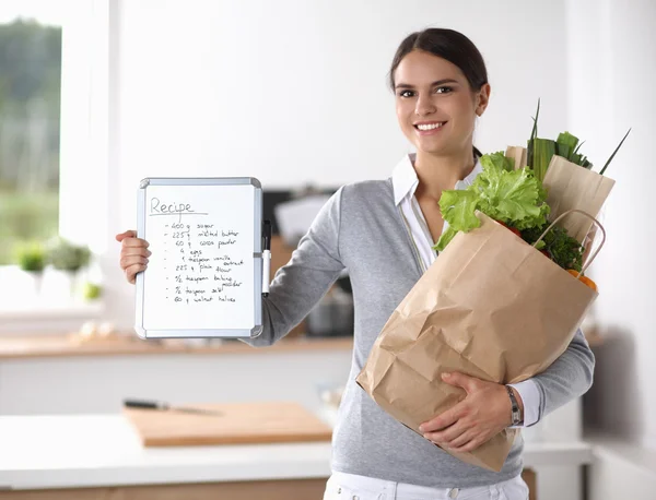 Mujer joven sosteniendo bolsa de la compra de comestibles con verduras de pie en la cocina. —  Fotos de Stock