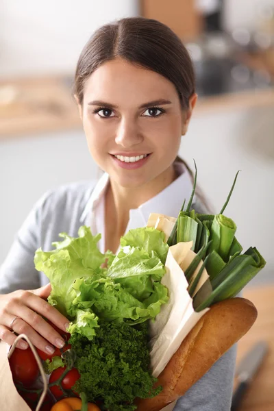 Mujer joven sosteniendo bolsa de la compra de comestibles con verduras de pie en la cocina. —  Fotos de Stock