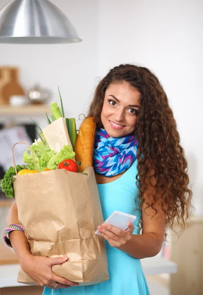 Mujer sonriente con teléfono móvil sosteniendo bolsa de compras en la cocina —  Fotos de Stock