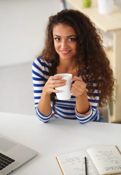 Lachende jonge vrouw met koffiekopje en laptop in de keuken thuis — Stockfoto