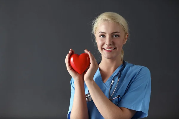 Doctor with stethoscope holding heart, standing on grey background — Stock Photo, Image