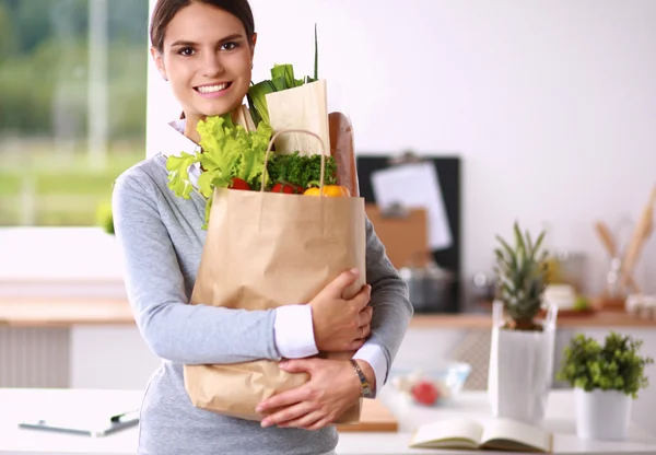 Mujer joven sosteniendo bolsa de la compra de comestibles con verduras de pie en la cocina. —  Fotos de Stock