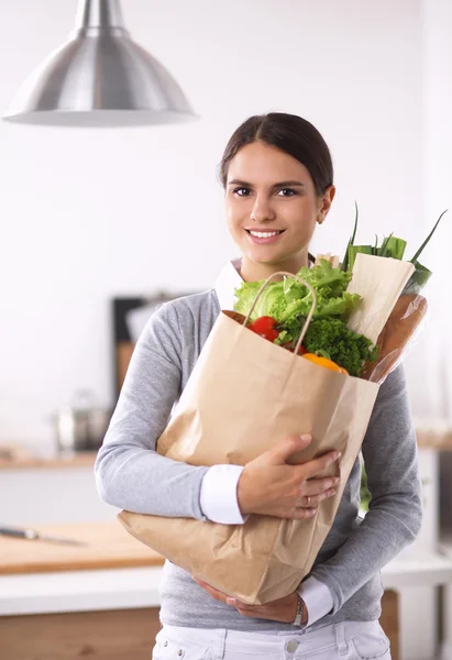 Mujer joven sosteniendo bolsa de la compra de comestibles con verduras de pie en la cocina. — Foto de Stock