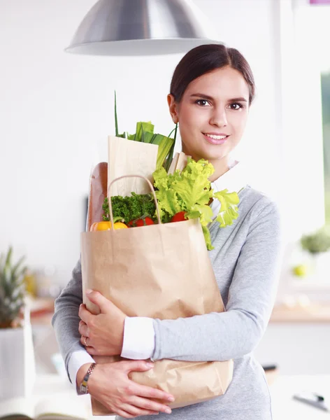 Mujer joven sosteniendo bolsa de la compra de comestibles con verduras de pie en la cocina. — Foto de Stock