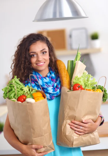 Mujer joven sosteniendo bolsa de la compra de comestibles con verduras de pie en la cocina. — Foto de Stock