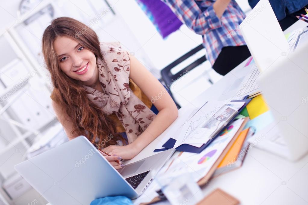 Young attractive female fashion designer working at office desk, drawing while talking on mobile