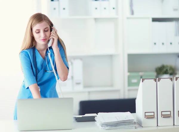 Jonge vrouw arts in witte jas aan de computer met behulp van telefoon — Stockfoto