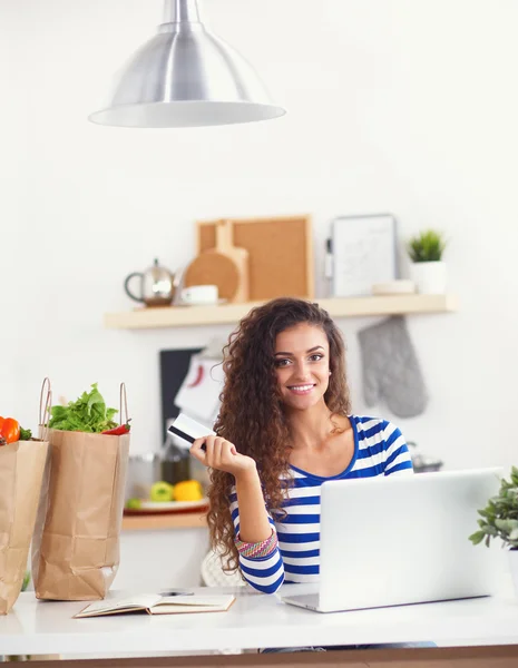 Smiling young woman with coffee cup and laptop in the kitchen at home — Stock Photo, Image