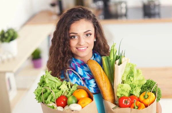 Mujer joven sosteniendo bolsa de la compra de comestibles con verduras de pie en la cocina. —  Fotos de Stock