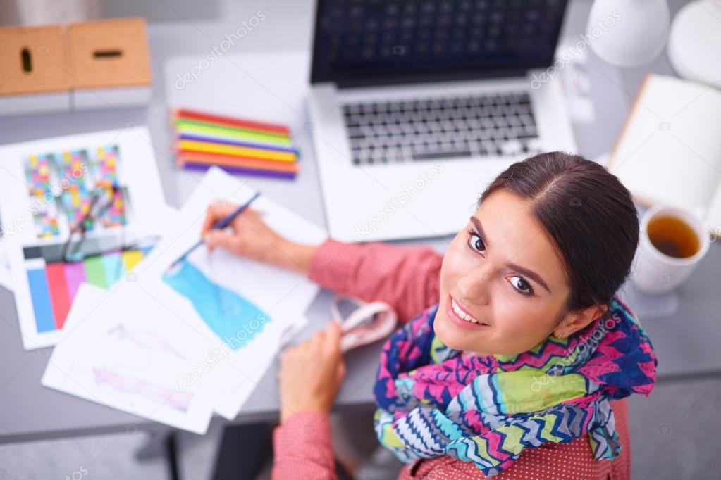 Young attractive female fashion designer working at office desk, drawing while talking on mobile