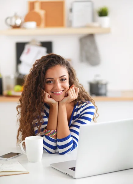 Smiling young woman with coffee cup and laptop in the kitchen at home — Stock Photo, Image