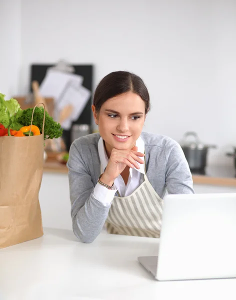 Mooie jonge vrouw koken op zoek naar laptop scherm met ontvangst in de keuken — Stockfoto