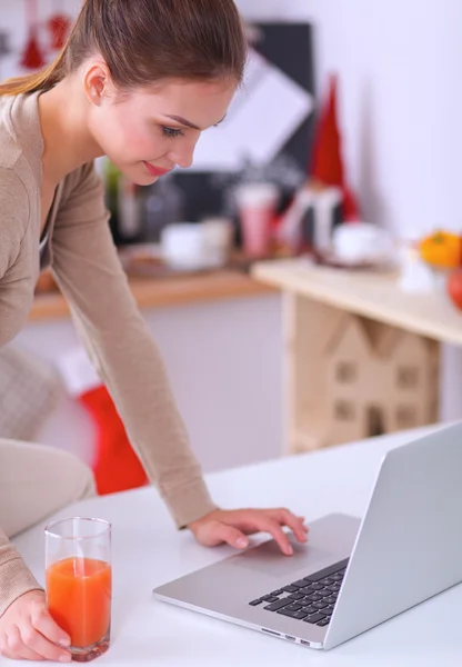 Woman using a laptop while drinking juice in her kitchen — Stock Photo, Image