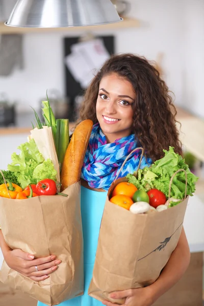 Mujer joven sosteniendo bolsa de la compra de comestibles con verduras. De pie en la cocina — Foto de Stock