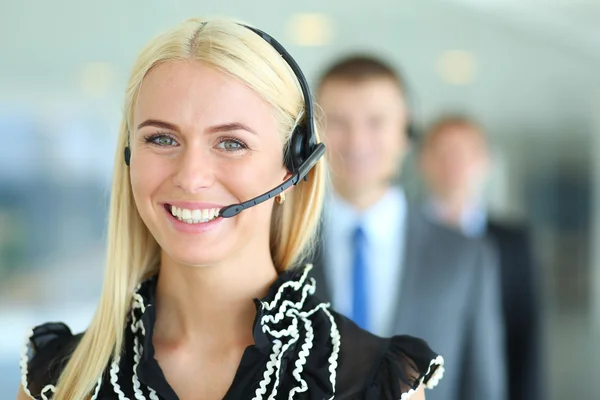 Mujer de negocios con auriculares sonriendo a la cámara en el centro de llamadas. Empresarios con auriculares en segundo plano —  Fotos de Stock