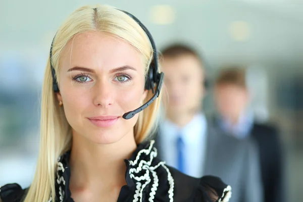Donna d'affari con auricolare sorridente alla telecamera in call center. Uomini d'affari in cuffia sullo sfondo — Foto Stock