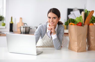 Güzel genç bayan yemek pişiriyor. Mutfakta makbuzu olan dizüstü bilgisayara bakıyor.