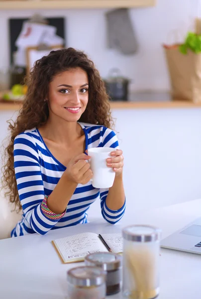 Smiling young woman with coffee cup and laptop in the kitchen at home — Stock Photo, Image