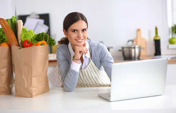 Mooie jonge vrouw koken op zoek naar laptop scherm met ontvangst in de keuken — Stockfoto
