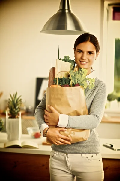 Mujer joven sosteniendo bolsa de la compra de comestibles con verduras de pie en la cocina. — Foto de Stock