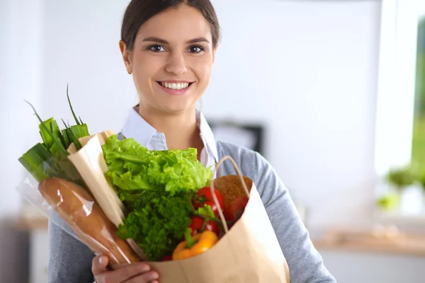 Mujer joven sosteniendo bolsa de la compra de comestibles con verduras de pie en la cocina. —  Fotos de Stock