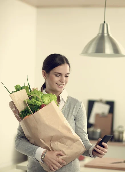 Mujer sonriente con teléfono móvil sosteniendo bolsa de compras en la cocina — Foto de Stock