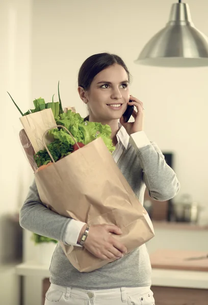 Mujer sonriente con teléfono móvil sosteniendo bolsa de compras en la cocina — Foto de Stock