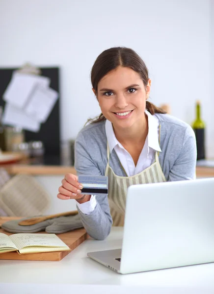 Mujer sonriente compras en línea utilizando la computadora y la tarjeta de crédito en la cocina —  Fotos de Stock