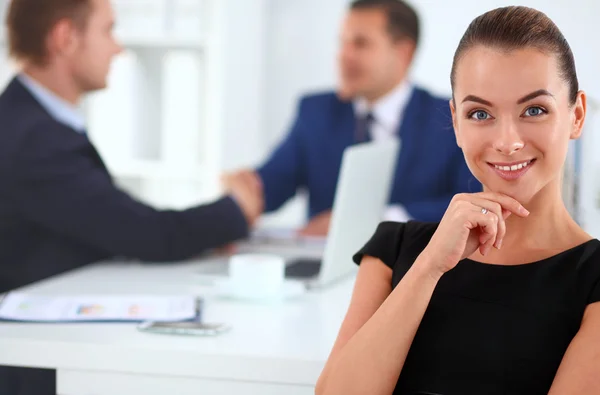 Portrait of a beautiful businesswoman sitting in an office with colleagues shaking hands in the background — Stock Photo, Image