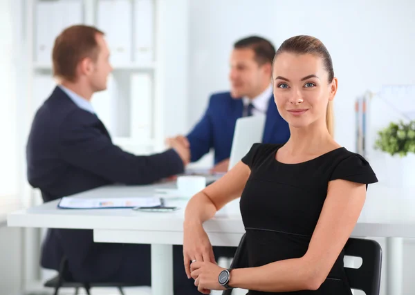 バック グラウンドで握手の同僚とオフィスで座っている美しい女性実業家の肖像画 — ストック写真