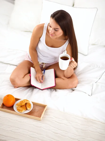 Unga vackra kvinnan sitta i sängen och skriva i ett häfte — Stockfoto