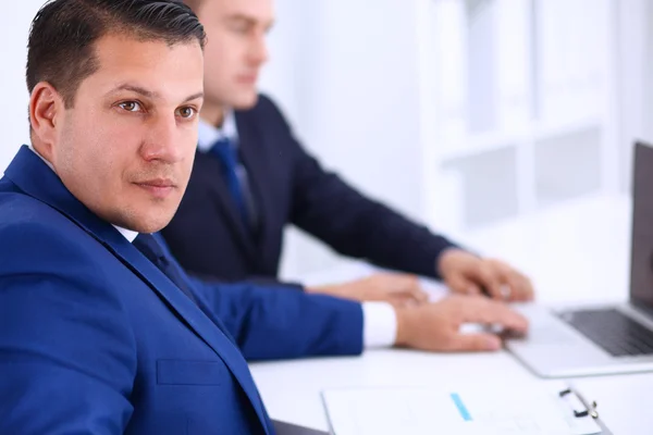 Porträtt av en stilig affärsman som sitter på ett kontor med kollegor i bakgrunden — Stockfoto