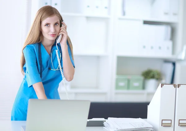 Jonge vrouw arts in witte jas aan de computer met behulp van telefoon — Stockfoto