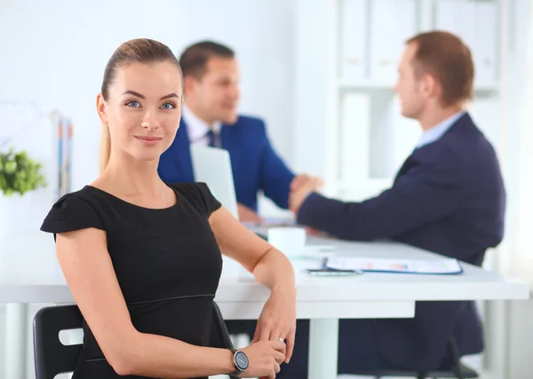 Retrato de uma bela mulher de negócios sentada em um escritório com colegas apertando as mãos no fundo — Fotografia de Stock