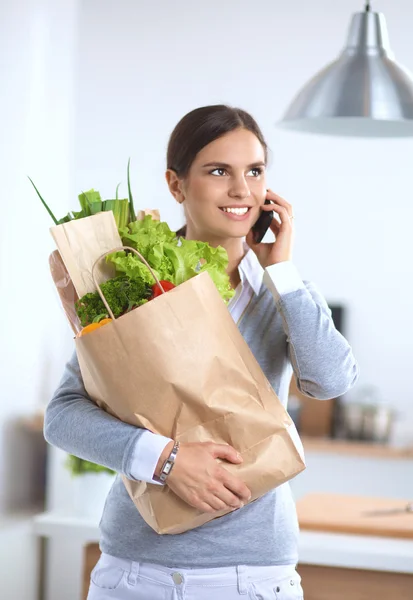 Usmívající se žena s mobilním telefonem držící nákupní tašku v kuchyni — Stock fotografie