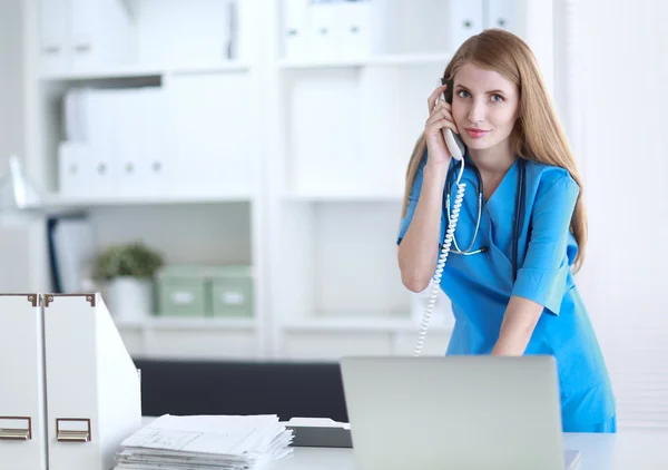 Mujer joven médico en bata blanca en el ordenador usando el teléfono — Foto de Stock