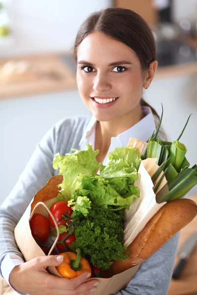 Mujer joven sosteniendo bolsa de la compra de comestibles con verduras de pie en la cocina — Foto de Stock