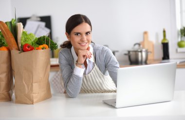 Güzel genç bayan yemek pişiriyor. Mutfakta makbuzu olan dizüstü bilgisayara bakıyor.