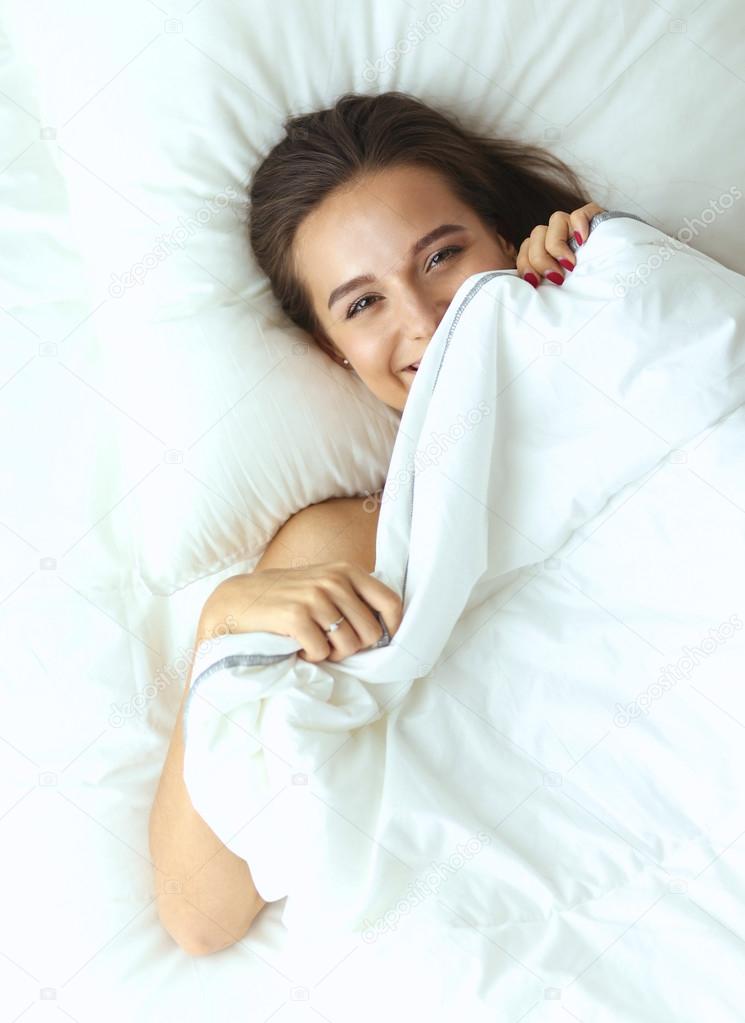 A pretty young woman peeking from under the covers in her bedroom happilly